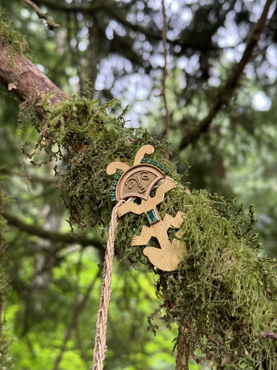A round gold amulet with green stones hanging on a moss-covered branch