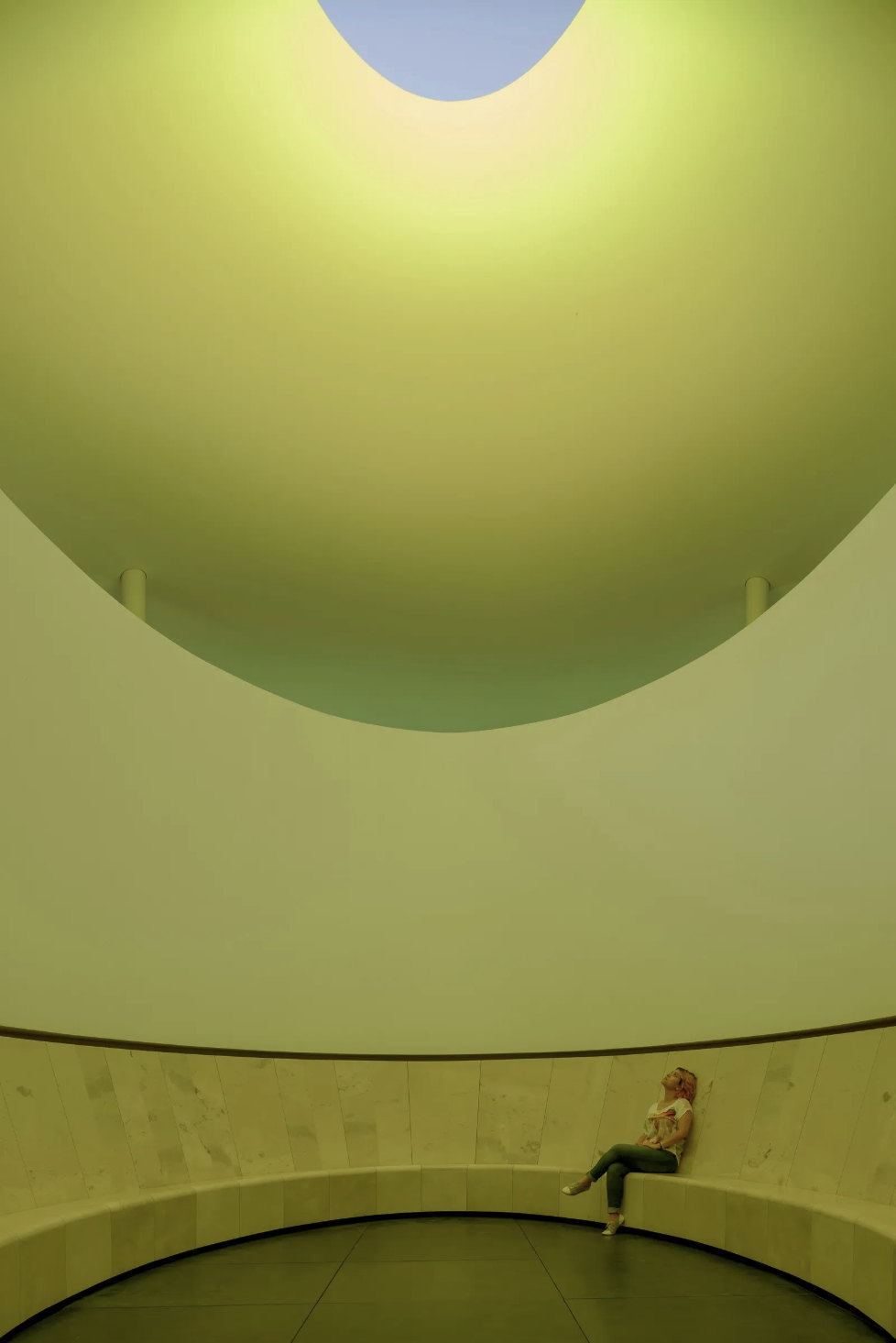 A woman seated alone on the bench of a round room, looking up through an opening in the ceiling to the sky